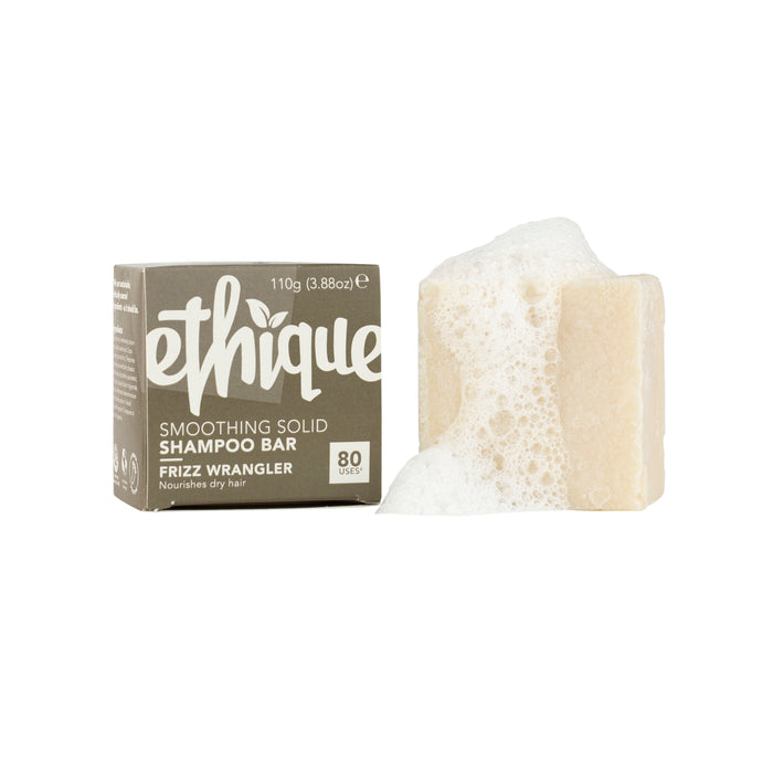 Ethiques Frizz Wrangler Smoothing Shampoo Bar gracefully rests next to its neutral packaging with white text, crowned with sudsy bubbles. Ideal for dry hair, it offers up to 80 uses and highlights its frizz-taming capabilities.