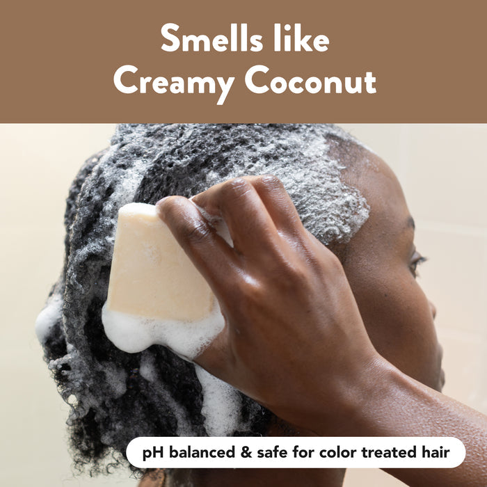 A person washing their curly hair with a bar of soap that is creating a rich lather. Text on the image reads, "Smells like Creamy Coconut." A smaller text at the bottom says, "pH balanced &amp; safe for color treated hair.