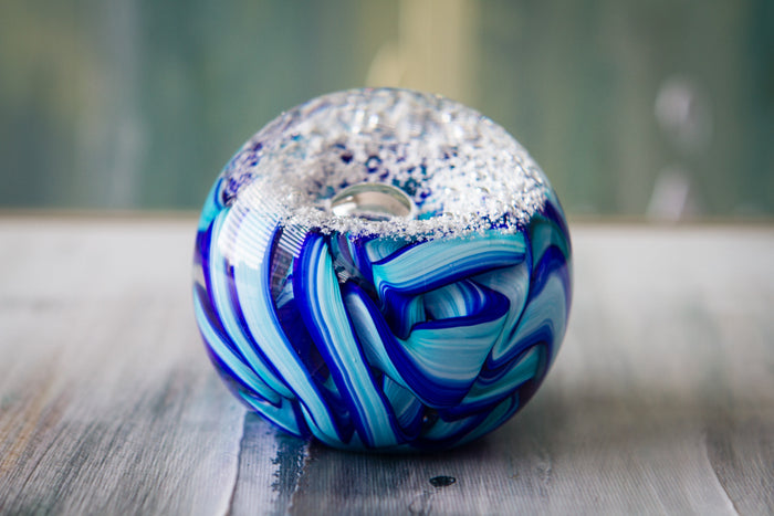 Blue glass memorial paperweight with teal and azure ribbon swirls and snowy top resting on a weathered wooden surface