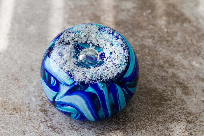 Round blue glass memorial paperweight with a snowy ring on top and bold blue ribbon patterns on a textured surface