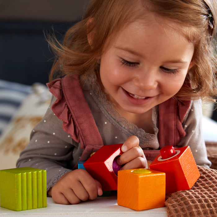 A young brown-haired child happily plays with HABAs eco-friendly Fun with Sounds - Wooden Discovery Blocks on a table. Wearing a pink dress over a gray shirt, she focuses on the colorful blocks, boosting her sensory development.