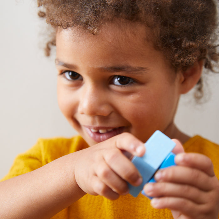 A young child with curly hair beams with joy, clutching a small blue block from their HABA Fun with Sounds - Wooden Discovery Blocks set, wearing a mustard yellow shirt. The blurred background highlights the childs cheerful and playful expression.