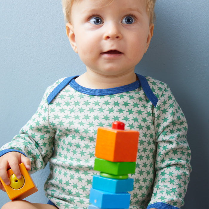 A toddler with light hair, wearing a star-patterned outfit, explores sensory development with HABAs Fun with Sounds - Wooden Discovery Blocks. Holding a yellow block, the child plays against a plain light blue wall backdrop.