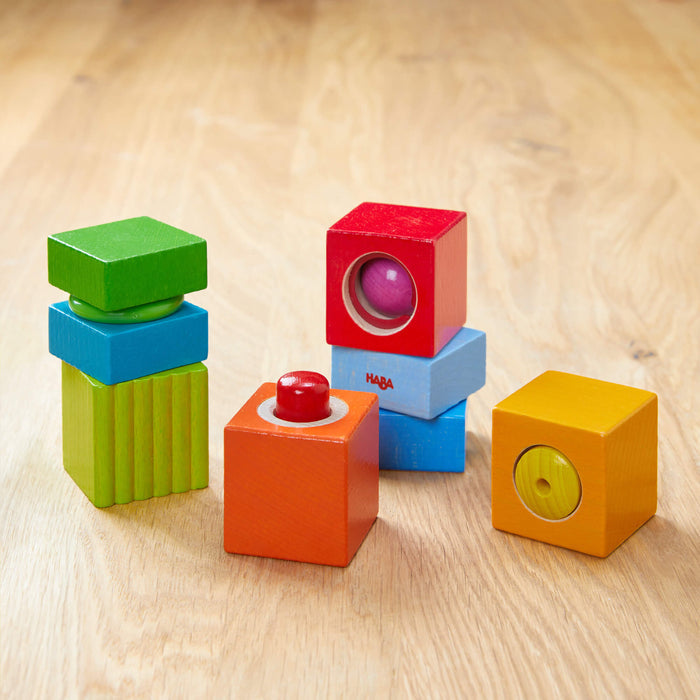 Fun with Sounds - Wooden Discovery Blocks by HABA are stacked on wood. The blocks, in vibrant red, green, blue, and orange, contain visible spheres for sensory development and are eco-friendly.