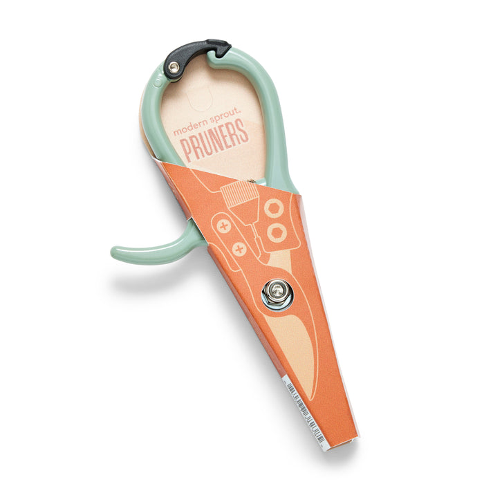 A pair of Modern Sprout Garden Pruners with a light green handle and durable high carbon steel blade is partially covered by an orange sheath featuring an illustration and the brand name.