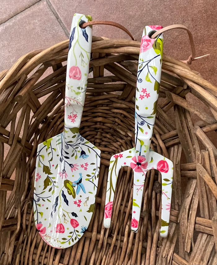 The Womanswork Garden Tool Set, essential for any gardener, includes a trowel and fork featuring the charming Garden of Paradise floral design with pink, green, and blue flowers and birds on white. Both tools have leather hanging loops and come in a woven basket.