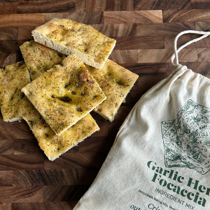 Slices of garlic herb focaccia lay on a wooden surface next to an opened cloth bag labeled FarmSteady Garlic Herb Focaccia Baking Mix, featuring a graphic of herbs and spices.