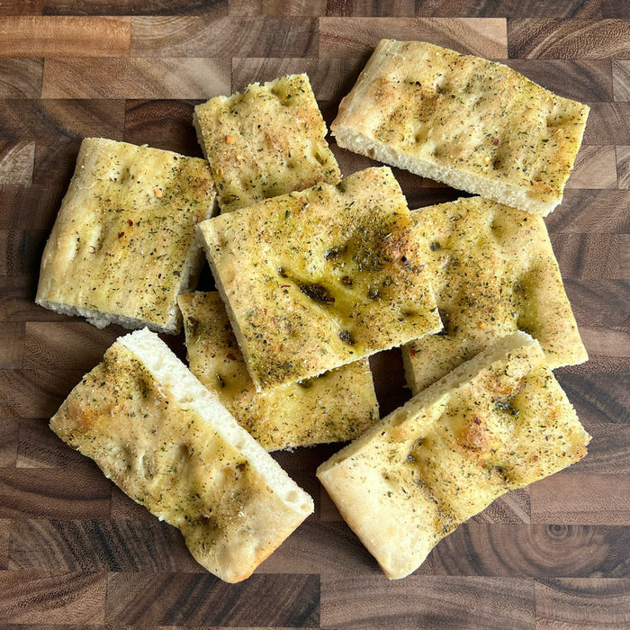 Slices of FarmSteadys Garlic Herb Focaccia are arranged on a wooden surface, featuring a golden-brown crust with visible rosemary and olive oil drizzles, showcasing the artistry possible with our baking mix.