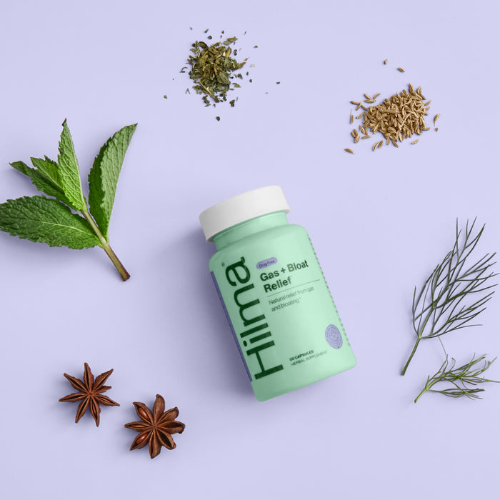 A green Hilma Gas + Bloat Relief bottle rests on a light purple surface, surrounded by mint leaves, fennel seeds, dried herbs, star anise, and fennel fronds—natural ingredients known for calming the digestive system.