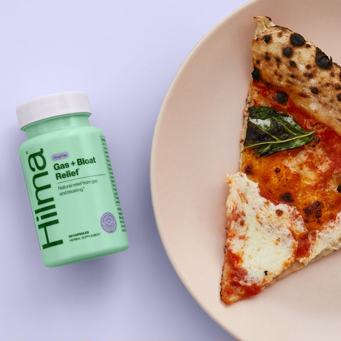 A bottle of Hilmas Gas + Bloat Relief, crafted with natural herbs to ease digestion, rests next to a cheesy pizza topped with tomato sauce and basil on a light purple surface.