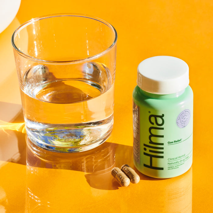 A clear glass of water and a green Hilma Gas + Bloat Relief bottle sit on a bright orange surface with two capsules, their shadows highlighting the promise of relief through natural herbs for digestive health.