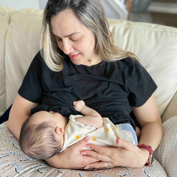 A woman in a black shirt sits on a light-colored couch, using the Haakaa Generation 1 Silicone Breast Pump with Lid while breastfeeding her baby. She gazes lovingly at the infant, dressed in white with yellow sunflowers, as she gently cradles them.