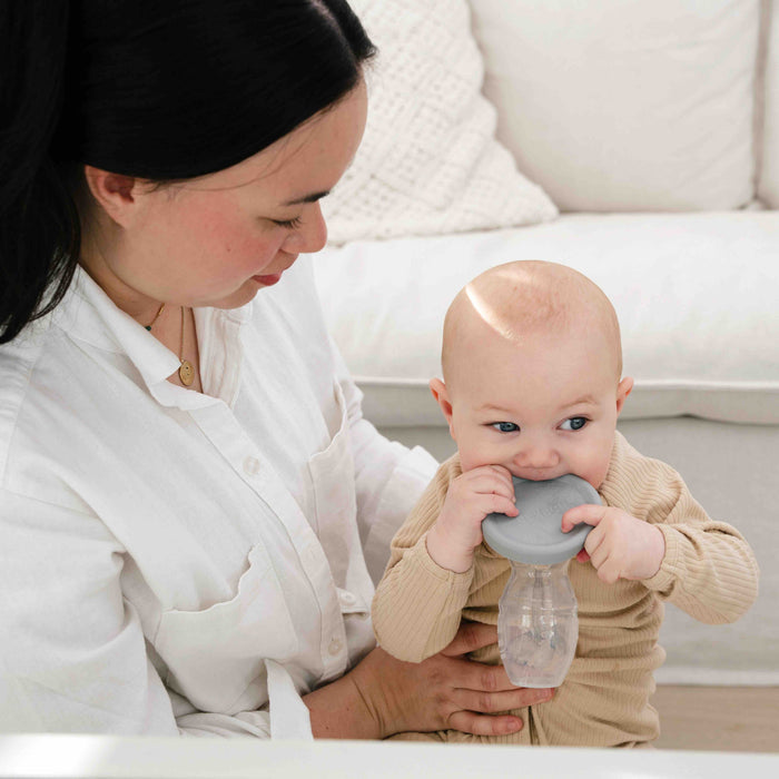 A dark-haired woman sits on a couch holding a baby and Haakaas Generation 1 Silicone Breast Pump with Lid. The baby, dressed in light clothes, chews on a gray object attached to the bottle. The cozy setting and pillows in the background suggest an eco-friendly lifestyle.