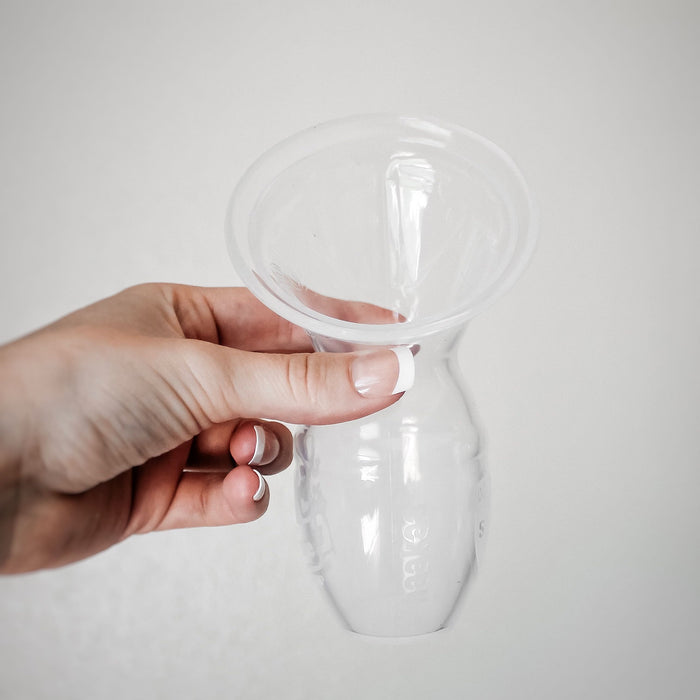 A hand holds a Haakaa Generation 1 Silicone Breast Pump with Lid against a plain light background. This clear, eco-friendly pump features a wide flared top and textured body for better grip. The persons neatly manicured nails are visible.