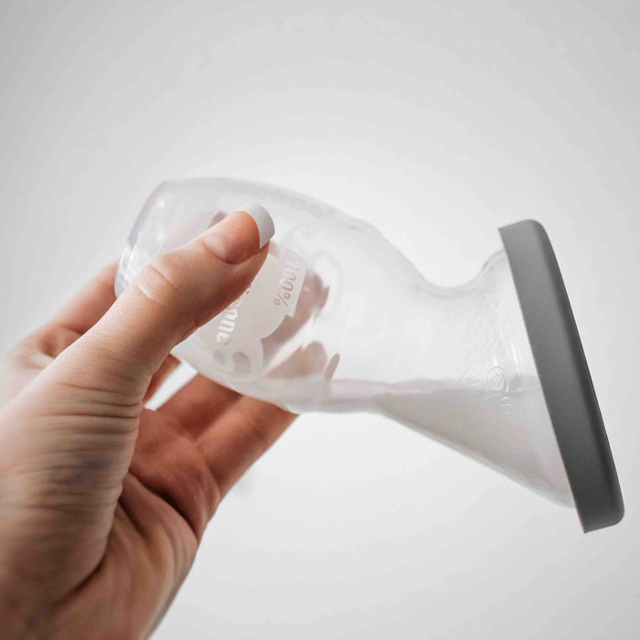 A hand gently holds the Haakaa Generation 1 Silicone Breast Pump with Lid, featuring a transparent silicone body and a gray rim, set against a light gray background.