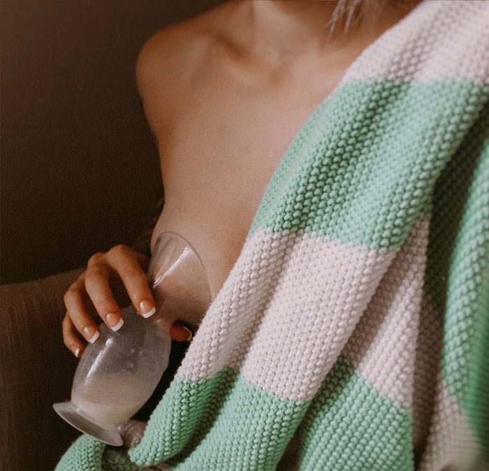 Partially covered with a green and white striped knitted blanket, a person uses the Haakaa Generation 2 Silicone Breast Pump with Lid, made from 100% food-grade silicone. Their hand holds the breast pump which has collected milk, creating a cozy and relaxed setting.