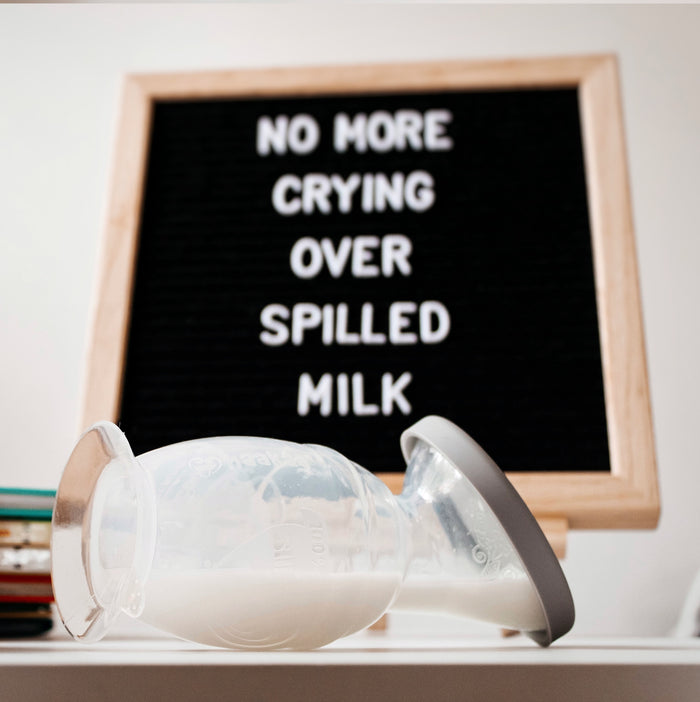A milk collection cup lies on its side with milk inside. In the background, a letter board reads "No more crying over spilled milk." The scene suggests resilience and humor in the face of minor mishaps.