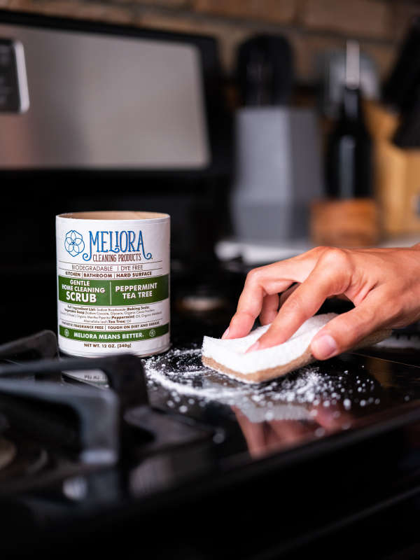 A hand cleans a black stovetop with Gentle Home Cleaning Scrub. A Meliora container labeled peppermint tea tree scented and septic-safe is nearby, surrounded by kitchen utensils, enhancing the eco-friendly cleaning ambiance.