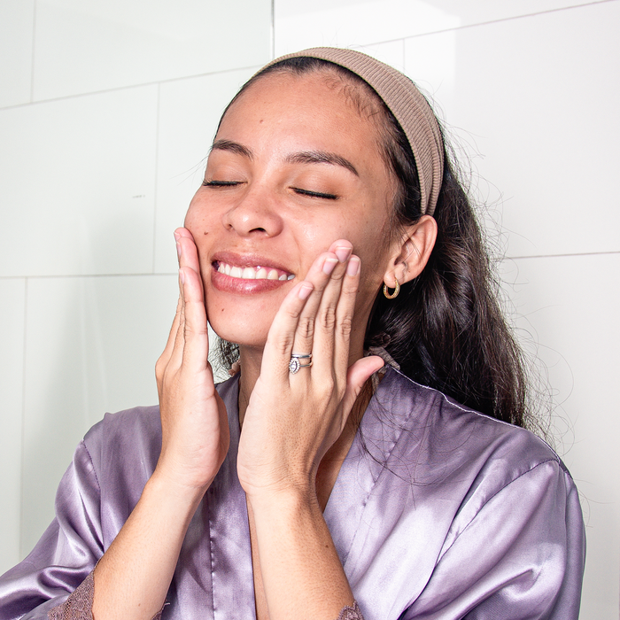 Wearing a headband and silk robe, a long-haired person smiles with closed eyes, touching their cheeks. Theyre enjoying the soothing effects of Tree To Tubs Gentle Retinol Alternative Bakuchiol Face Serum for Smooth and Radiant Skin, indulging in their skincare routine.
