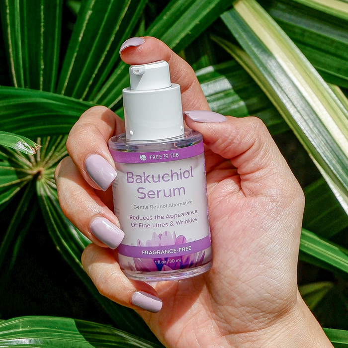 A hand with light purple nails holds a bottle of Tree To Tubs Gentle Retinol Alternative Bakuchiol Face Serum for Smooth and Radiant Skin against green leaves, highlighting its gentle efficacy in reducing fine lines and wrinkles.