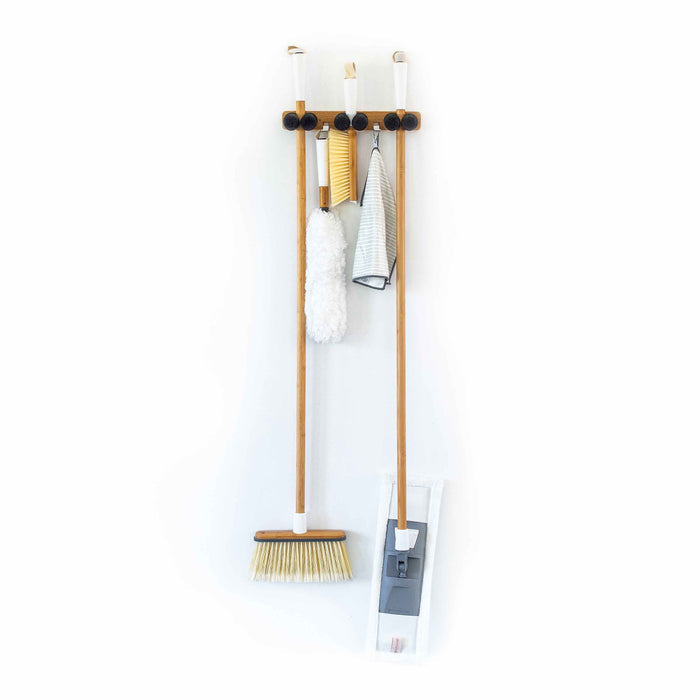 The Get A Grip - Mop & Broom Organizer by Full Circle mounts on a plain white wall, efficiently storing wooden-handled brooms and mops, along with cleaning tools like a duster and cloths held in clips above.