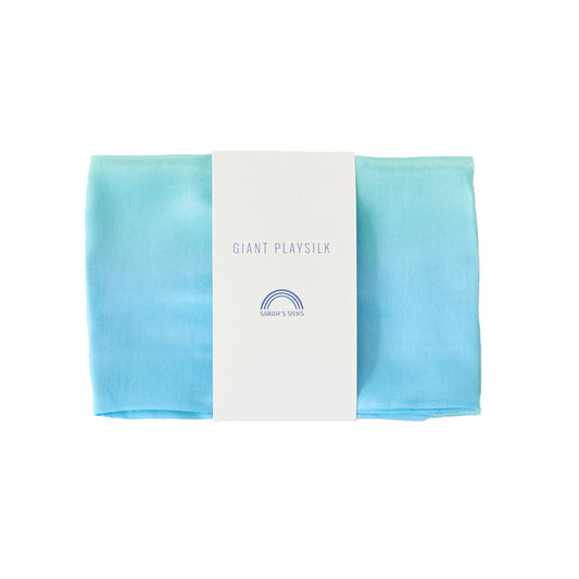 The Giant Playsilk by Sarahs Silks, ideal for open-ended play in Montessori and Waldorf homes, displays a gradient from light blue to turquoise. It is wrapped with a white paper band labeled Giant Play Silk featuring a small rainbow graphic.
