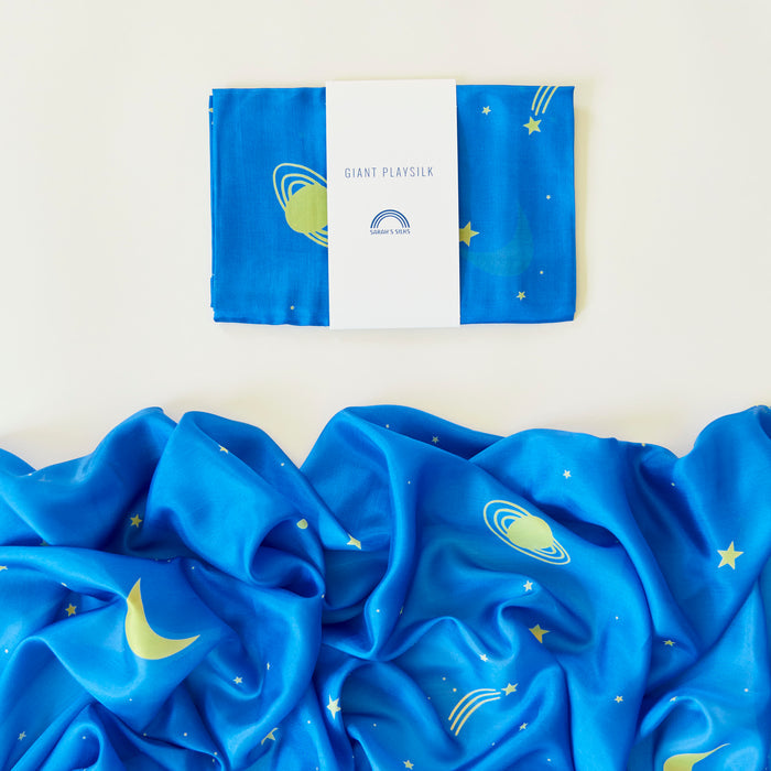 A playsilk from Sarahs Silks, called Giant Playsilk for Open-Ended, Imaginative Play, features a blue space theme with planets, crescents, stars, and swirls. Ideal for Montessori & Waldorf homes, it rests elegantly on a light background.