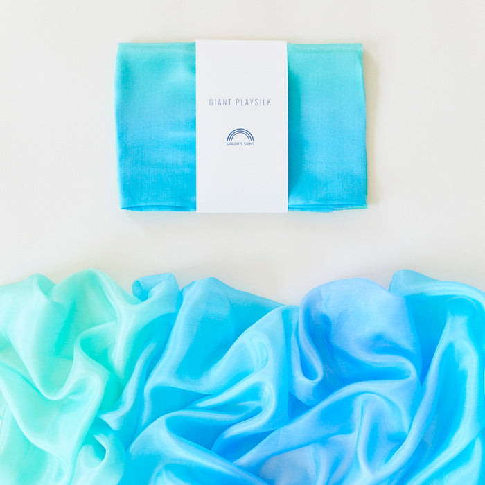 A folded turquoise Giant Playsilk by Sarahs Silks, featuring a small rainbow icon on the label, rests above an unfolded wavy silk in shades of blue and turquoise against a white background—ideal for open-ended play in Montessori & Waldorf homes.