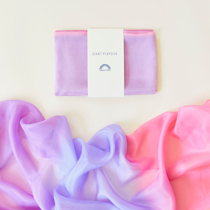 Folded and packaged under the Giant Playsilk for Open-Ended, Imaginative Play by Sarahs Silks, the purple silk with a small rainbow logo suits Montessori & Waldorf homes. Nearby lies an unfolded pink and purple piece on a flat surface, ready for open-ended play.
