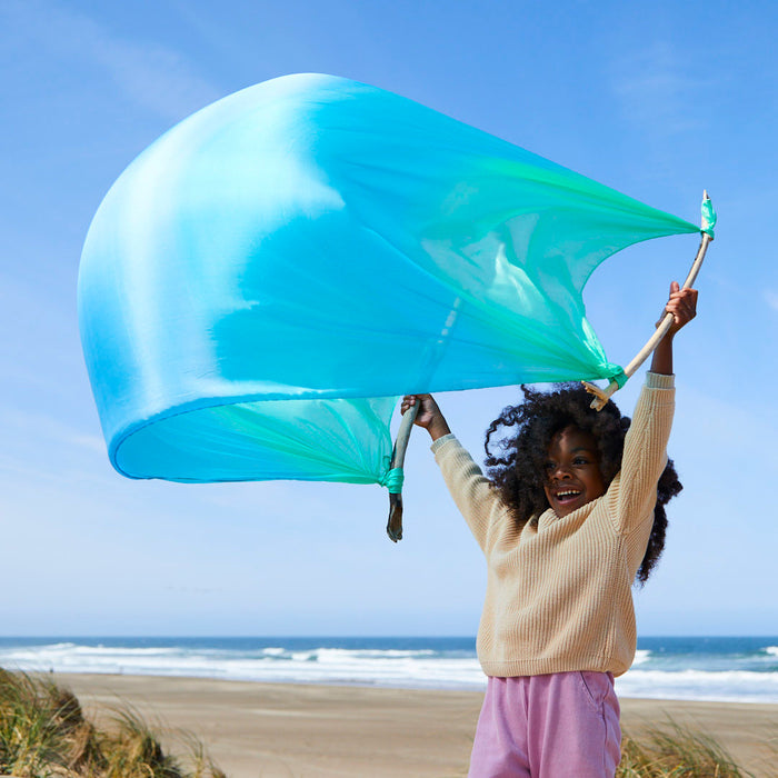 A child joyfully plays on a sandy beach with Sarahs Silks Giant Playsilk, embracing open-ended imaginative play. The large blue and green fabric dances against the bright sky and ocean backdrop, evoking the creativity found in Montessori & Waldorf homes.