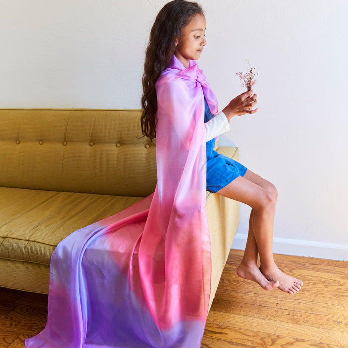 A young girl with long hair sits on a yellow couch, wrapped in Sarahs Silks Giant Playsilk for Open-Ended, Imaginative Play in pink and purple. Holding dried flowers and wearing a white and blue outfit, she gazes thoughtfully, embodying open-ended play.