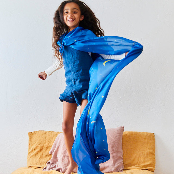 In her Montessori-inspired room, a joyful young girl with long, wavy hair jumps on her bed wearing blue overalls and waving a Giant Playsilk from Sarahs Silks. The scarf is star and moon-decorated, complementing the mustard yellow and soft pink pillows that invite imaginative play adventures.
