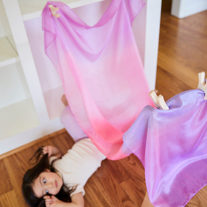A young child lies on a wooden floor surrounded by Sarahs Silks Giant Playsilks in soft pink and purple, draped over a low shelf and secured with clothespins. The shelves are empty as the child gazes calmly upward, enjoying their open-ended, imaginative play.