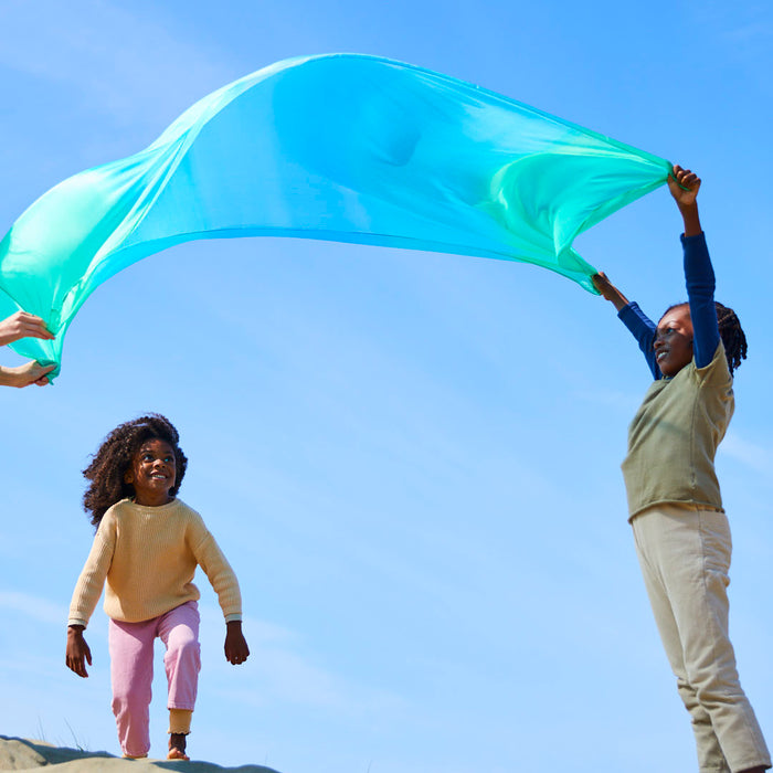 Two children enjoy open-ended play with a blue-green Giant Playsilk from Sarahs Silks, flowing against the clear sky. One lifts it high as the other runs beneath on a sandy hill, embracing creativity cherished in Montessori and Waldorf homes on this sunny day.