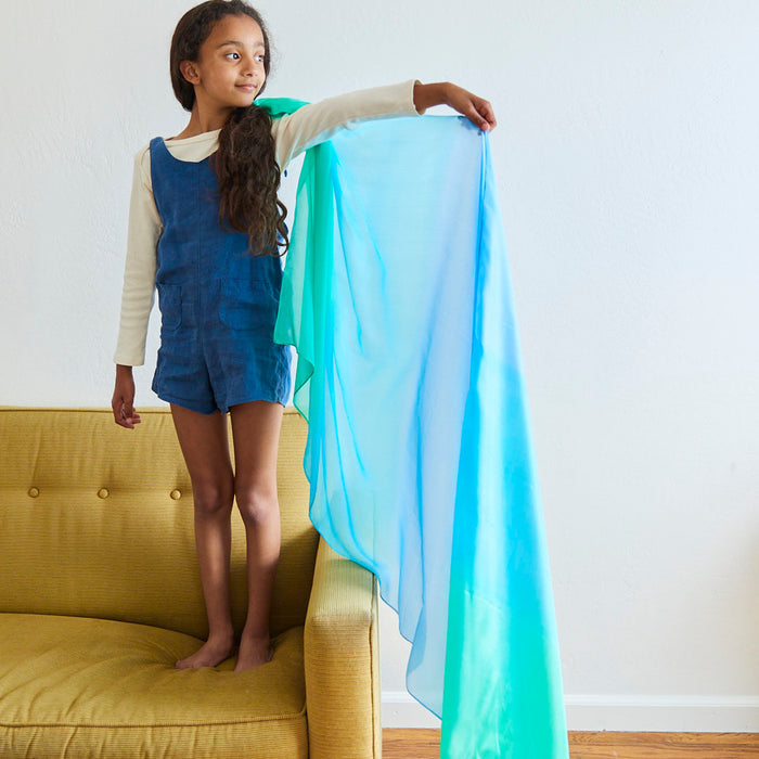 A young girl with long hair stands on a yellow sofa, holding up a flowing blue and green Sarahs Silks Giant Playsilk for Open-Ended, Imaginative Play like a cape. She wears a blue dress over a long-sleeve shirt and gazes into the distance in her well-lit Montessori & Waldorf home.