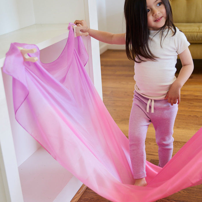 A young girl with long dark hair, in a white shirt and purple pants, joyfully holds Sarahs Silks Giant Playsilk for Open-Ended, Imaginative Play attached to a shelf in a Montessori & Waldorf-style room with wooden floors and a sofa.