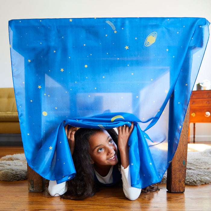 A child with long hair peeks out from under a table covered with Sarahs Silks Giant Playsilk, featuring stars and planets—ideal for open-ended play. The room, modeled after Montessori & Waldorf styles, includes a wooden floor, fluffy rug, yellow sofa, and wooden cabinet in the background.