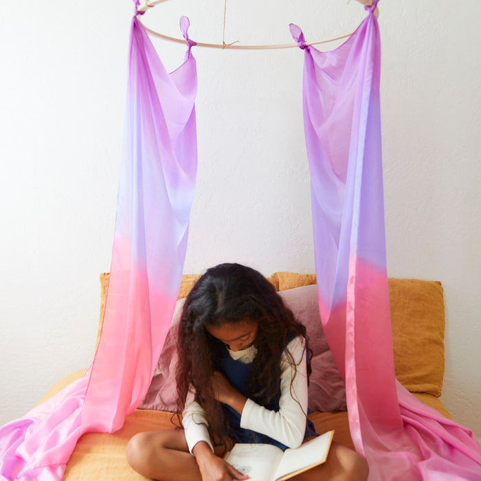 A child with long hair sits cross-legged on a bed, reading a book. Pink and purple Giant Playsilks from Sarahs Silks hang above, forming a whimsical canopy reminiscent of Montessori and Waldorf homes. The backdrop is a simple white wall with mustard-colored bedding.