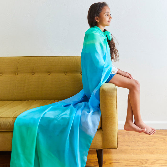 A young girl with long hair sits barefoot on a yellow couch armrest, wearing the Giant Playsilk for Open-Ended, Imaginative Play by Sarahs Silks. The blue and green cape drapes elegantly to the wooden floor, embodying Montessori and Waldorf homes spirit of open-ended play.