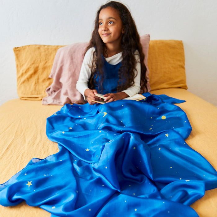 A girl sits on a bed with a yellow blanket, holding a book. A blue blanket with star and moon patterns, similar to the Giant Playsilk by Sarahs Silks, is draped over her legs. She smiles at a pink pillow.