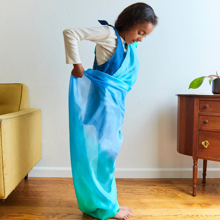 A child stands barefoot on a wooden floor, wrapped in a flowing blue and green Giant Playsilk from Sarahs Silks. Smiling, they embrace imaginative play. Nearby, a yellow chair and small wooden table with a potted plant create a cozy Montessori & Waldorf-inspired space.
