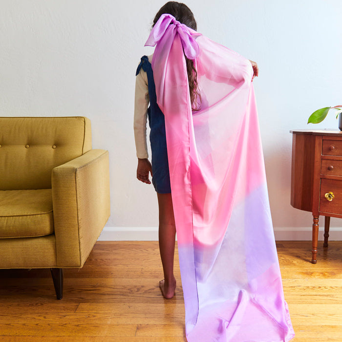 In a Montessori-style living room, a child with long hair, dressed in blue, transforms using Sarahs Silks Giant Playsilk in pink and purple as a cape. A yellow sofa complements the wooden side table with a plant, creating the perfect backdrop for open-ended play.