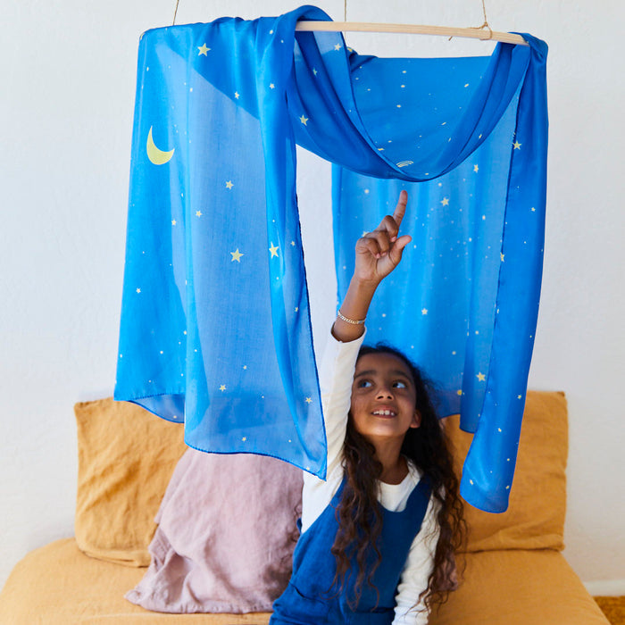 A child pointing up from a sofa under a blue, star-patterned canopy captures the playful charm of Montessori & Waldorf homes. Surrounded by mustard and pink pillows against a white wall, this setting exudes the whimsical appeal of Sarahs Silks Giant Playsilk for open-ended, imaginative play.