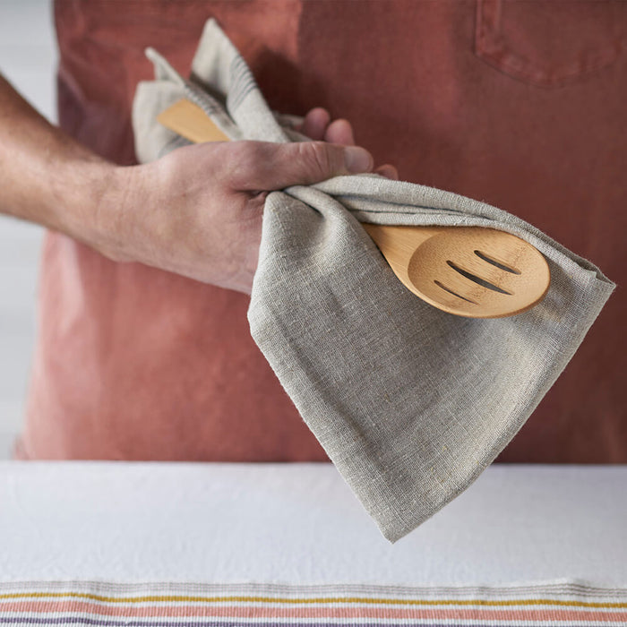 A person in a reddish apron holds a Give it a Rest Slotted Spoon by Bambu, wrapped in a beige cloth napkin, standing before a table covered with a white tablecloth with subtle striped edging.