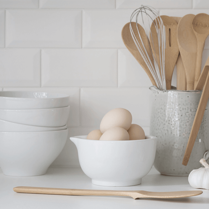 On a white kitchen counter, a bowl of eggs, stacked bowls, and various utensils including a whisk and wooden spoons are in a speckled ceramic holder. A Give it a Rest Spatula by Bambu rests near some garlic, enhancing the display of organic kitchen tools.