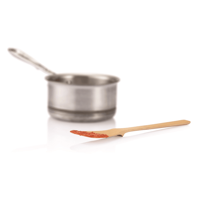 A stainless steel saucepan with a long handle is in the background. In the foreground, Bambus Give it a Rest Spatula, made of organic bamboo, holds a small amount of tomato sauce on its edge against a white, reflective surface.