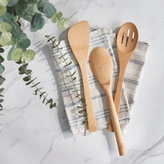 The Bambu Give it a Rest Spatula rests on a folded, striped cloth napkin atop a marble surface, with eucalyptus branches nearby.