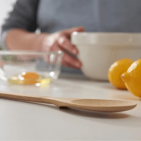 A Bambu 'Give It a Rest' Bamboo Spoon rests on a white countertop near two lemons, a bowl with a cracked egg, and someone preparing ingredients in the background.