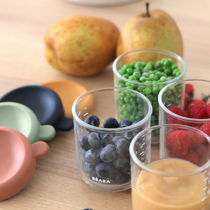Four BEABA Glass Baby Food Jars filled with blueberries, green peas, raspberries, and applesauce sit on a wooden table, colorful storage lids nearby and two pears in the background.
