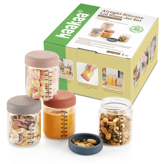 Three Haakaa glass jars with silicone lids, ideal for reusable glass container needs, hold marshmallows, sauce, and mixed nuts. Situated before packaging labeled Glass Baby Food Storage Jar Set, the jars include measurement markings and usage tips for homemade meals.